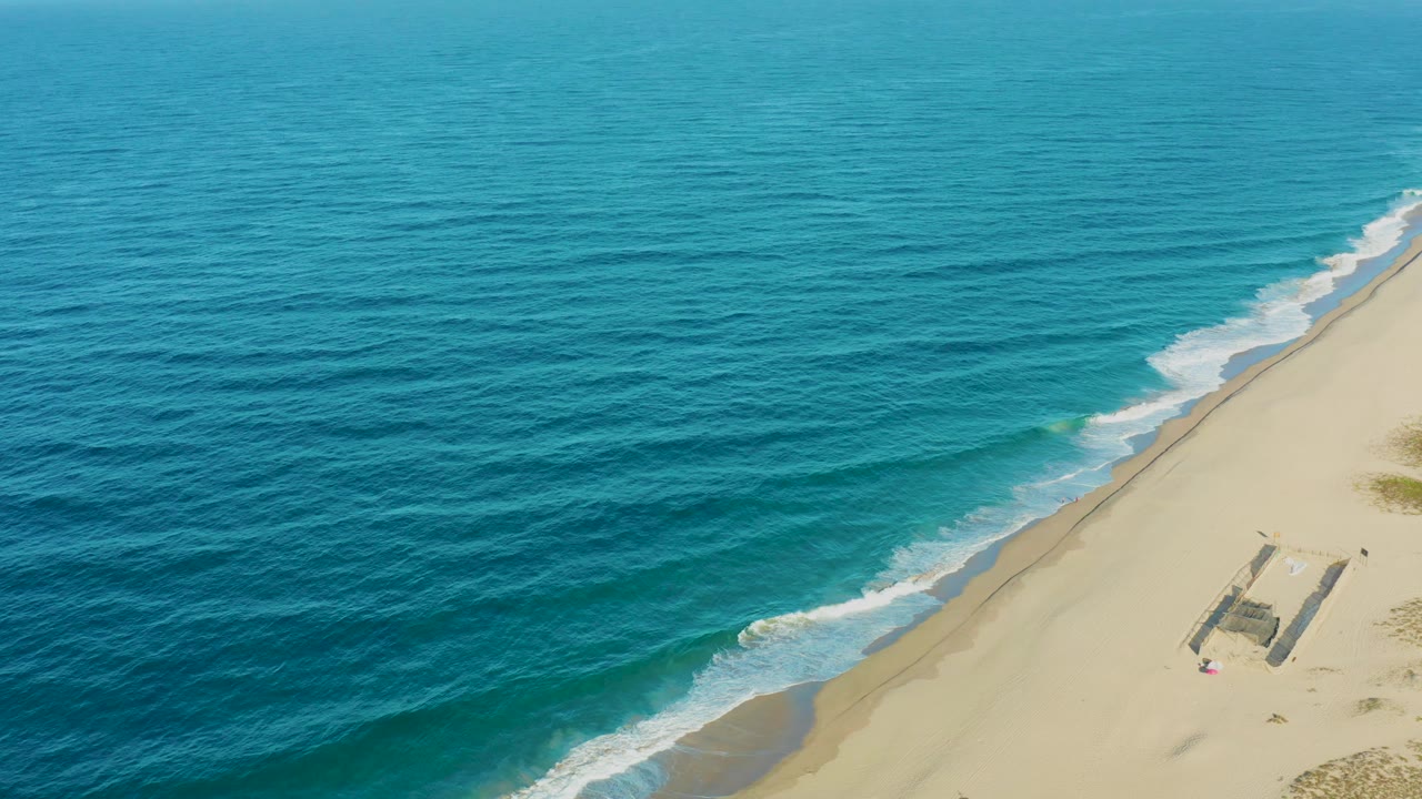 Blue ocean and sandy shoreline seen from above with surf breaking beside a small beach structure.