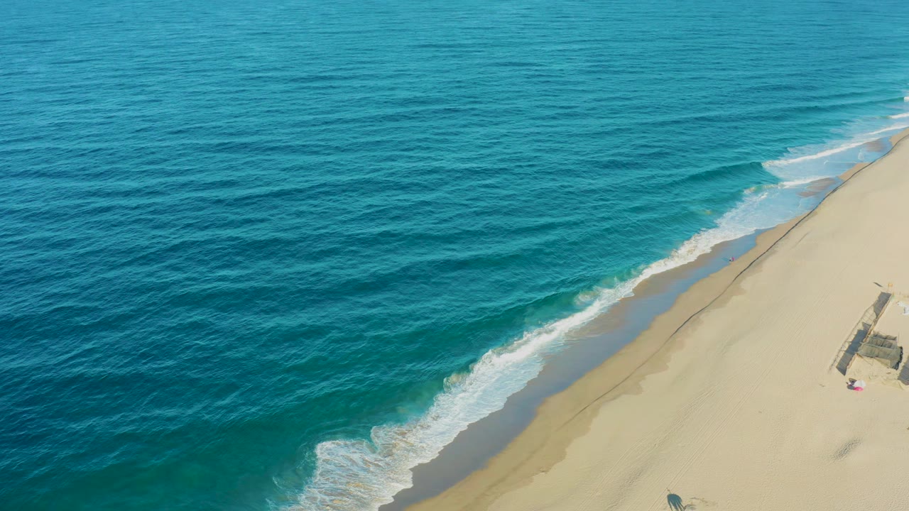 Aerial frame over Todos Santos with deeper blue-green water at left, white breakers at the shore, and empty sand along the right side, no visible people.
