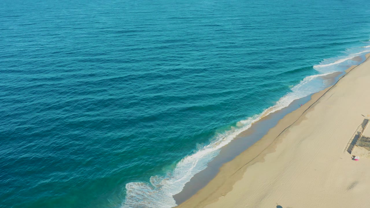 From an elevated view over Todos Santos, gentle surf approaches the pale beach in a long diagonal line, with the frame free of visible people.