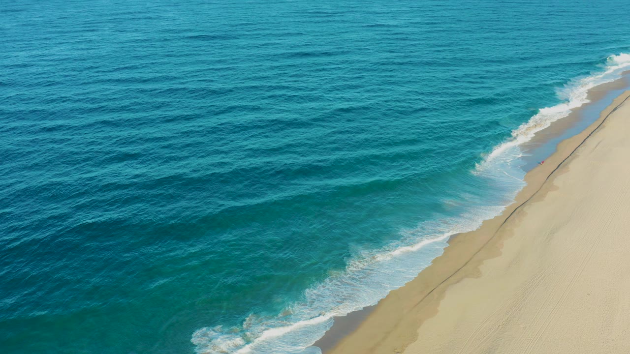 Overhead view in Todos Santos, Baja California Sur, Mexico, with blue ocean on the left, a diagonal sandy shoreline on the right, and no visible people.