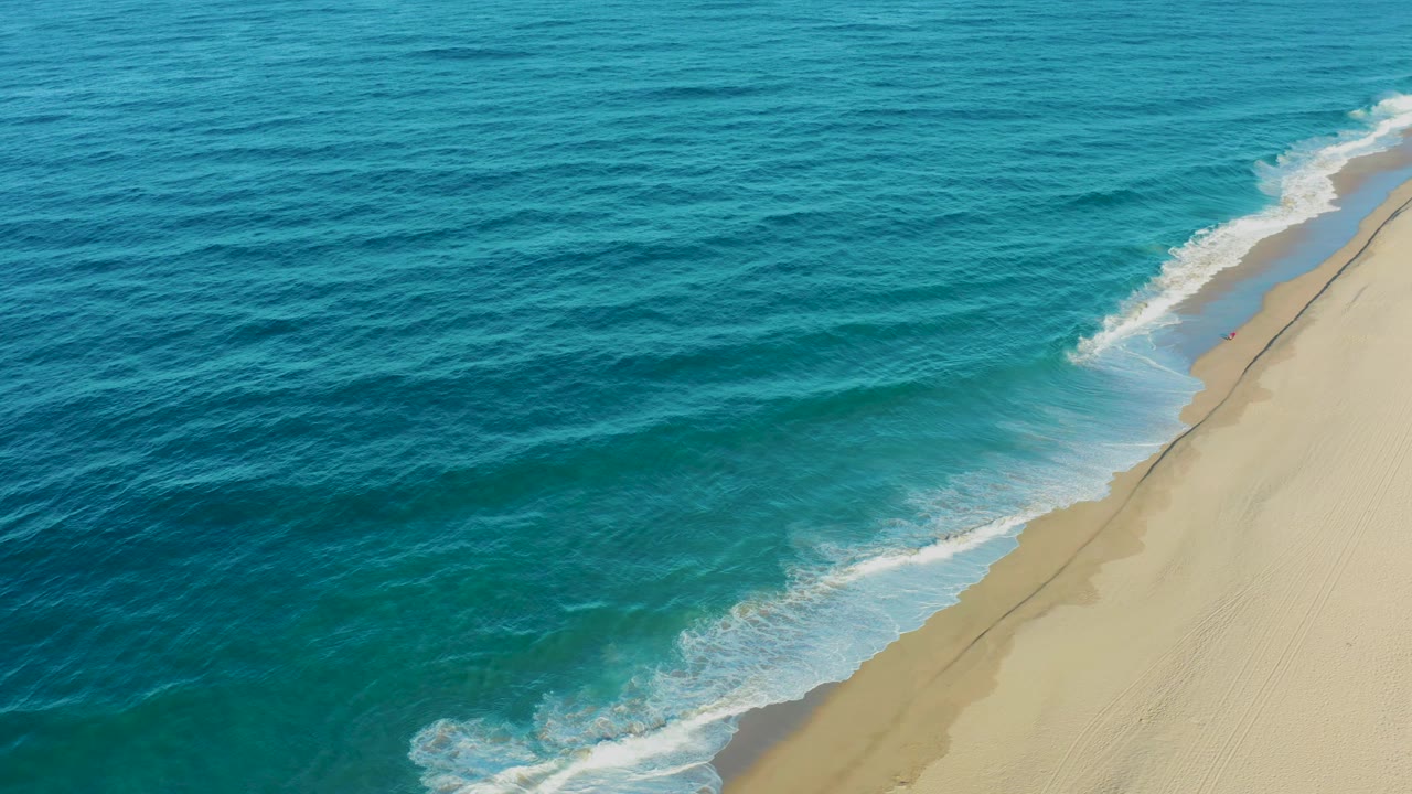 Todos Santos aerial view with darker blue water offshore, lighter turquoise water near the beach, a thin foam line, and no visible people.
