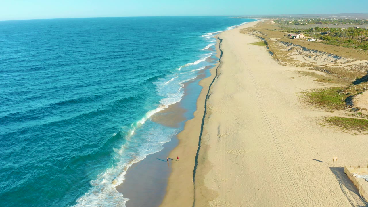 Aerial view over Todos Santos, Baja California Sur, with turquoise ocean, white surf, a broad sandy beach, dunes, distant buildings, and a small beachgoer near the waterline.