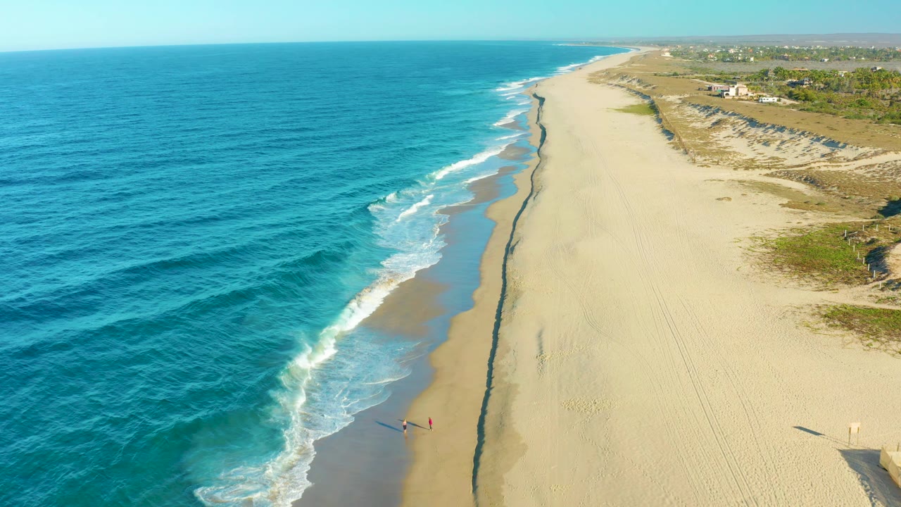 Drone-style aerial over Todos Santos coastline with deeper blue water filling the left side, surf curling onto the sand, dunes to the right, and a tiny beachgoer still visible near shore.