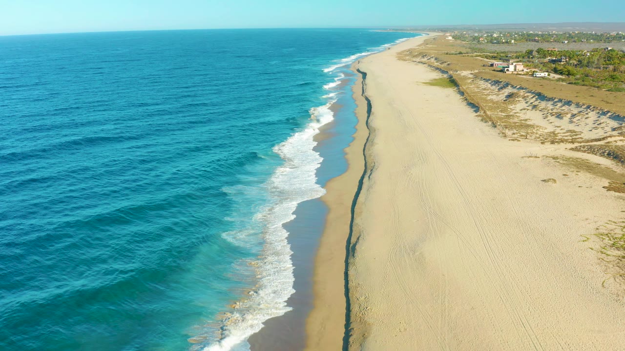 Aerial view over Todos Santos coastline with turquoise water, a curling band of white waves, pale sand, low dunes, sparse vegetation, and no visible people.