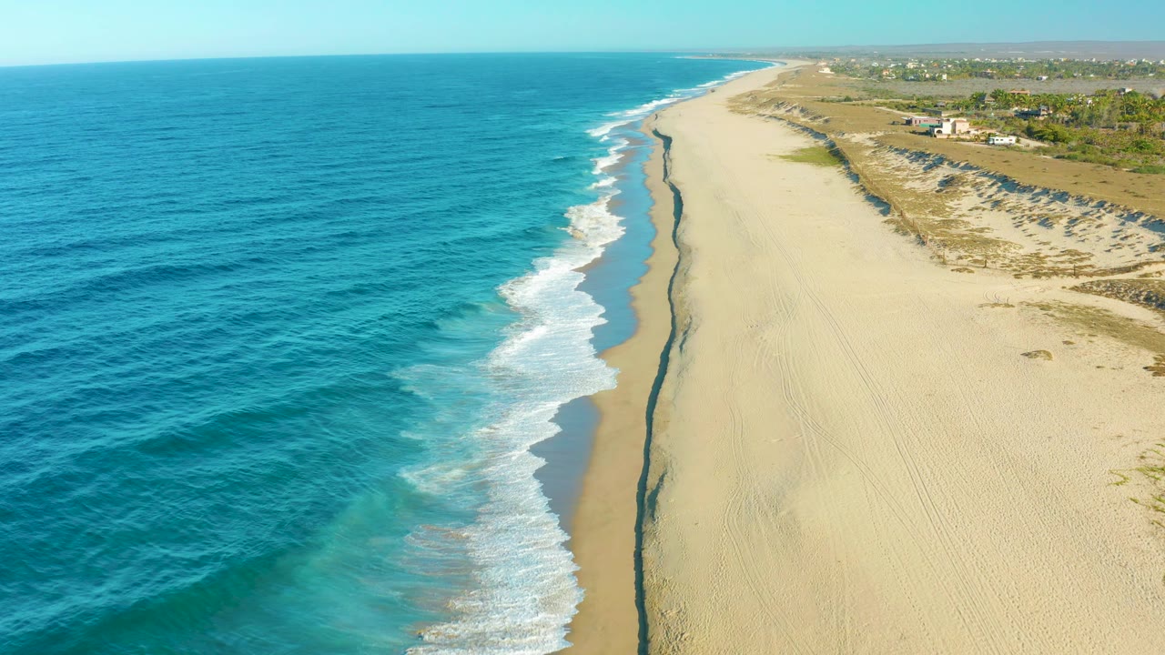 Todos Santos aerial frame with deeper blue water at left, foamy waves meeting the shore, a long pale beach, dune vegetation, small distant buildings, and no visible people.