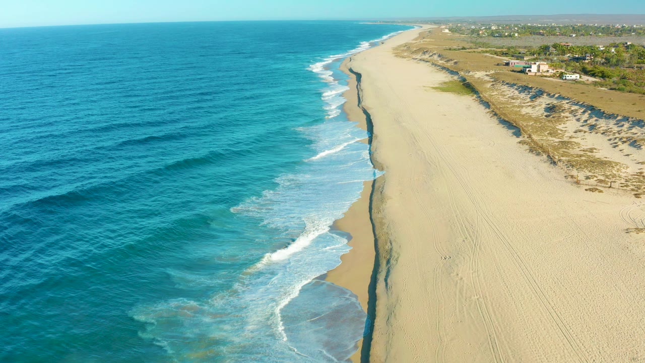Aerial view over Todos Santos, Baja California Sur, with deep blue ocean on the left, white surf meeting a wide sandy beach, low dunes and distant structures to the right, and no visible people.
