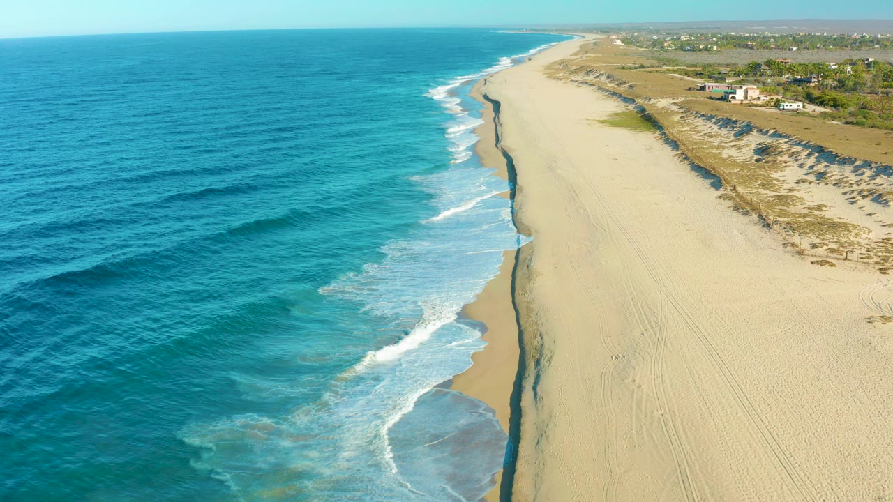 Elevated Todos Santos shoreline view with turquoise water shifting to darker blue offshore, thin bands of white foam on the sand, dune vegetation inland, and no visible people.