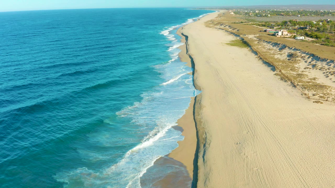 Todos Santos aerial beach frame with a larger band of foamy surf near center, pale sand stretching up the right side, darker ocean texture offshore, distant buildings beyond the dunes, and no visible people.