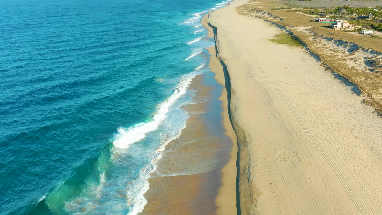 Aerial frame over a Todos Santos, Baja California Sur beach with turquoise water on the left, white surf crossing wet sand, dry tracked sand and dunes on the right, and no visible people.