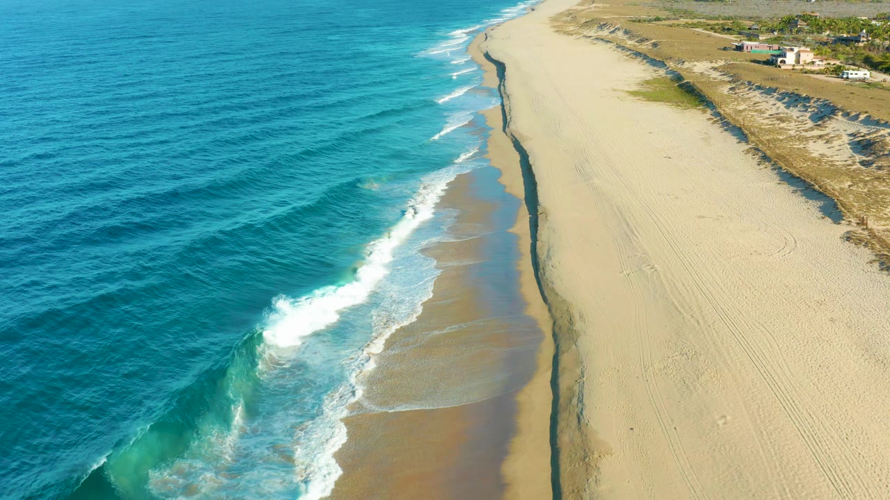 At 0:01, foamy waves push farther up the wet shoreline below the aerial view, with blue ocean to the left, open sand and dunes to the right, and no visible people.