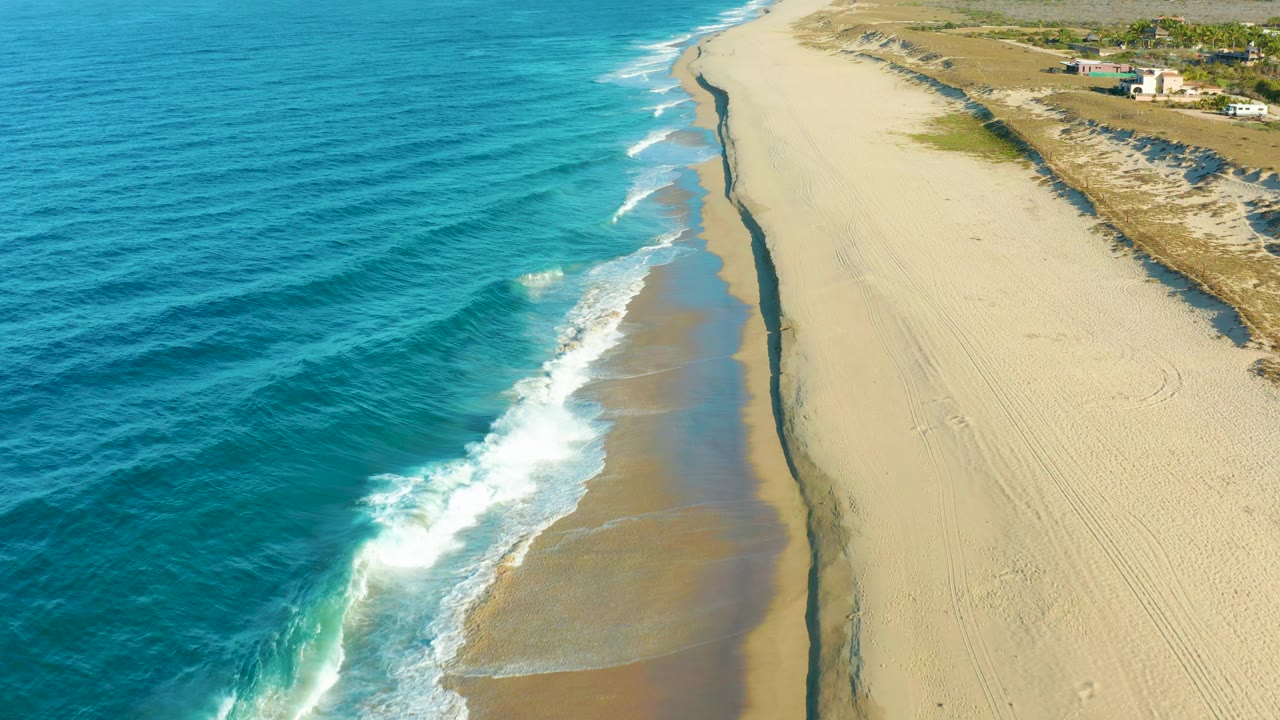 At 0:02, a bright band of breaking surf sits near the center of the Todos Santos beach view, with textured ocean water left, pale sand right, distant buildings inland, and no visible people.