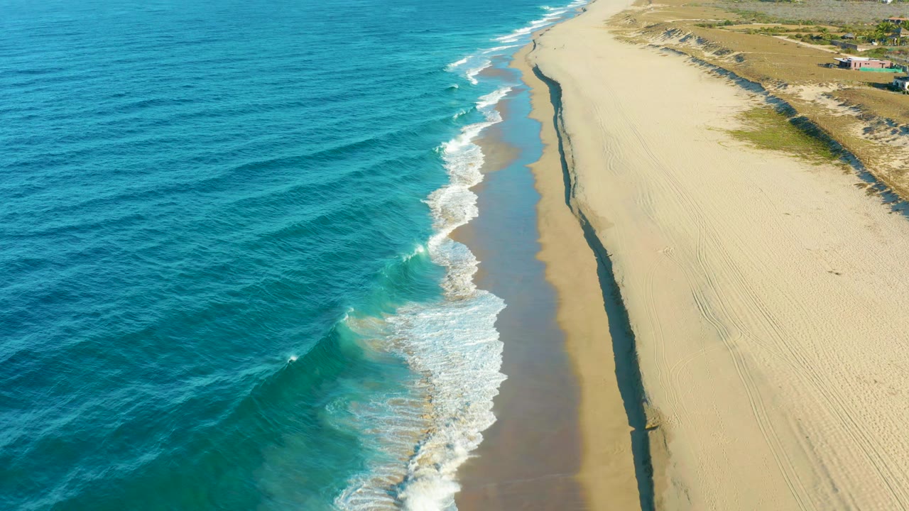Aerial frame of Todos Santos surf curling toward the beach, with a darker wet strip beside pale sand and no visible people on the shore.