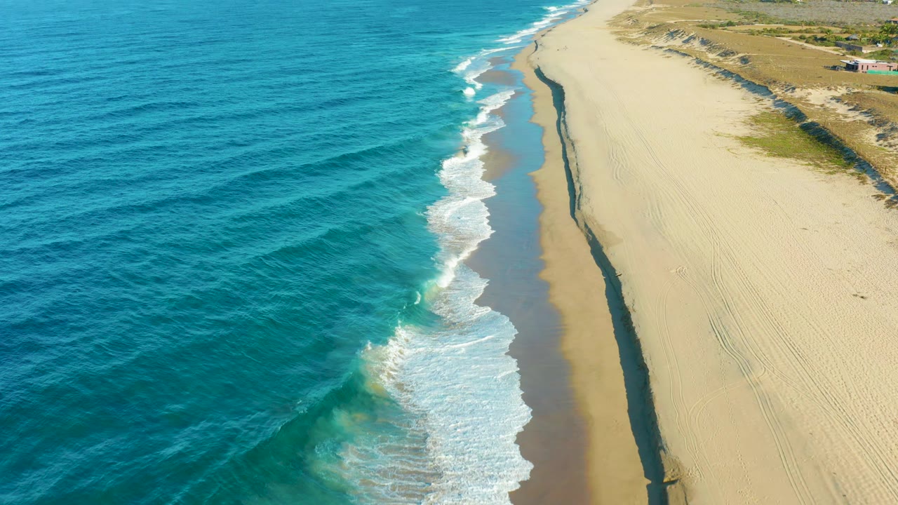 Todos Santos shoreline seen from above, where turquoise water darkens offshore, white foam spreads onto the sand, and no visible people appear.