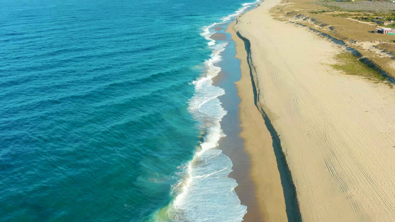 Elevated view of the Todos Santos coast as a wave sheet spreads across the lower beach, with blue water left, open sand right, and no visible people.