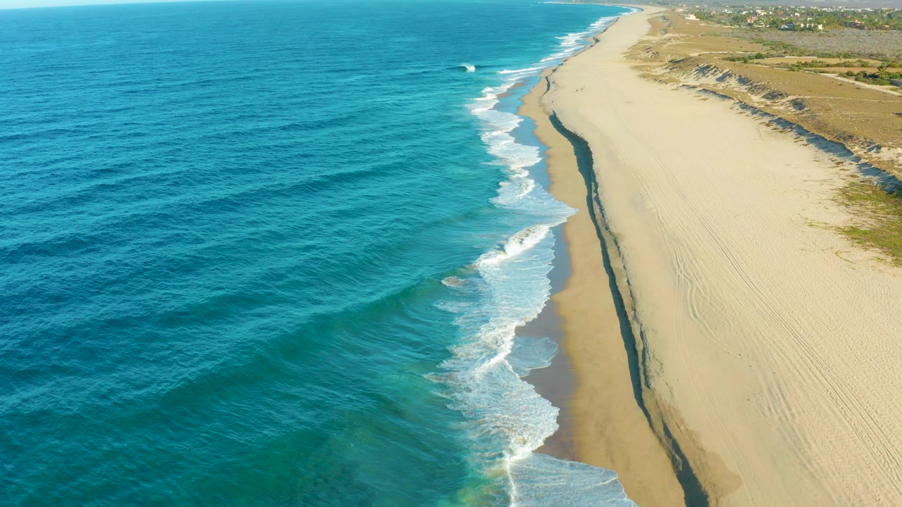 Opening aerial frame over Todos Santos, Baja California Sur, with blue ocean on the left, white surf tracing the sandy beach, dunes on the right, distant buildings near the horizon, and no visible people.