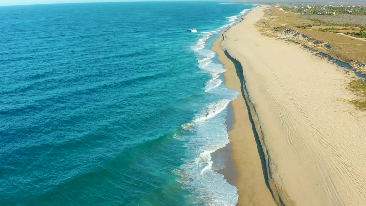 Aerial coastal frame in Todos Santos with darker blue ocean swell filling the left, bright foam breaking along the beach center, dune scrub on the right edge, and no visible people.