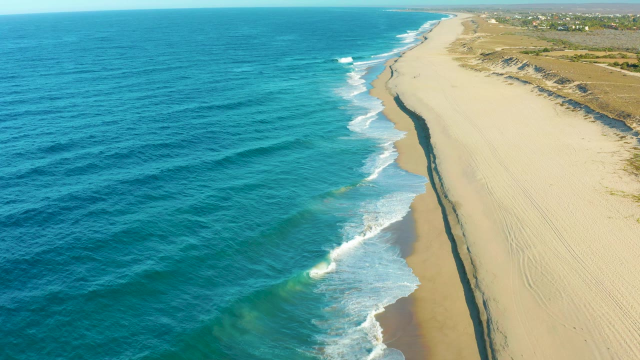 Aerial beach view with rolling waves, sand, dunes, and blue horizon.