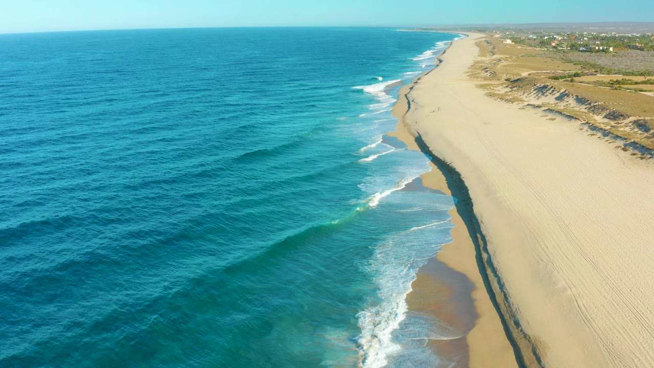 Aerial view in Todos Santos, Baja California Sur, with deep blue ocean at left, a diagonal line of white surf, pale sand at right, distant dunes, and no visible people.