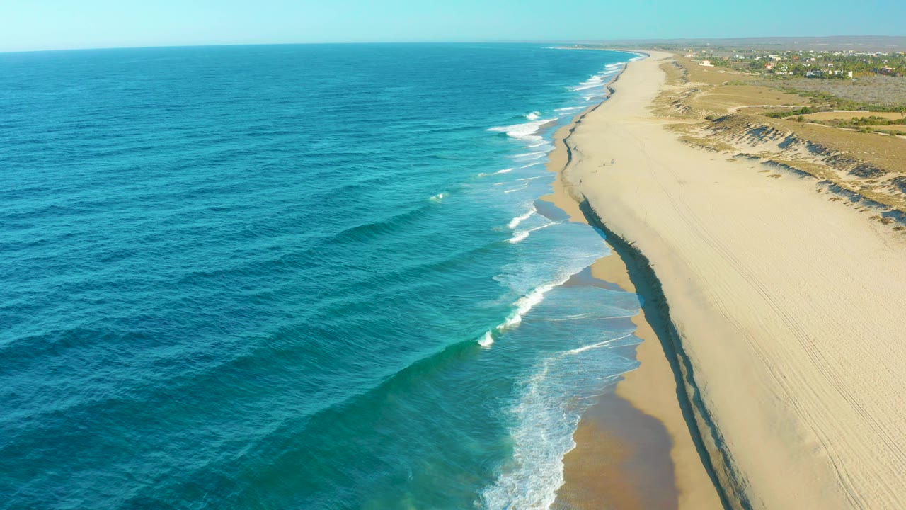 Elevated Todos Santos beach view with broad pale sand on the right, layered blue water on the left, a narrow foam line at the shoreline, distant inland structures, and no visible people.