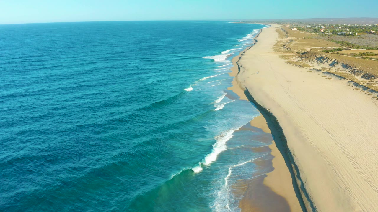 Todos Santos aerial coastline with open ocean occupying the left half, small wave sets rolling onto the sand, low dune ridges inland, a distant horizon, and no visible people.