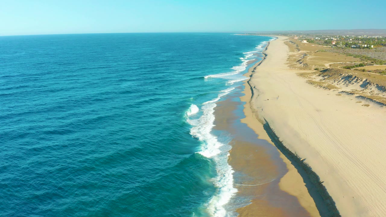 Aerial view over the Todos Santos coastline with blue ocean on the left, white surf on wet sand, low dunes on the right, and no visible people.