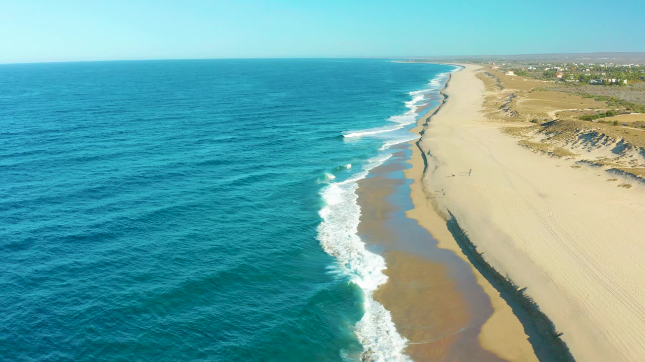 Aerial view of the Todos Santos shore with turquoise ocean taking more of the frame, white breakers curling toward the beach, dunes to the right, and no visible people.