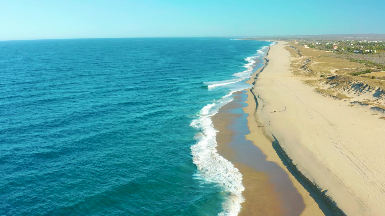 Aerial view of surf meeting a sandy coast with ocean to the left and dunes to the right.