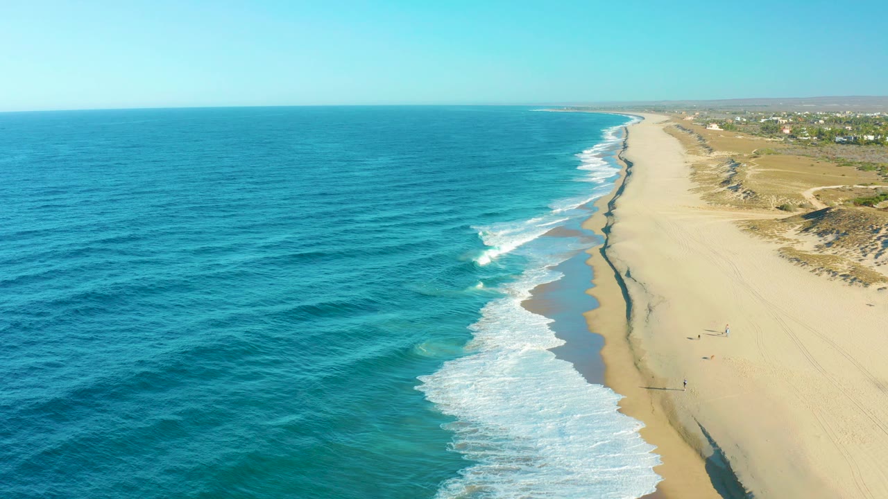 Aerial view of Todos Santos coastline with turquoise ocean on the left, white surf reaching a broad sandy beach, dunes and small distant buildings in the background.