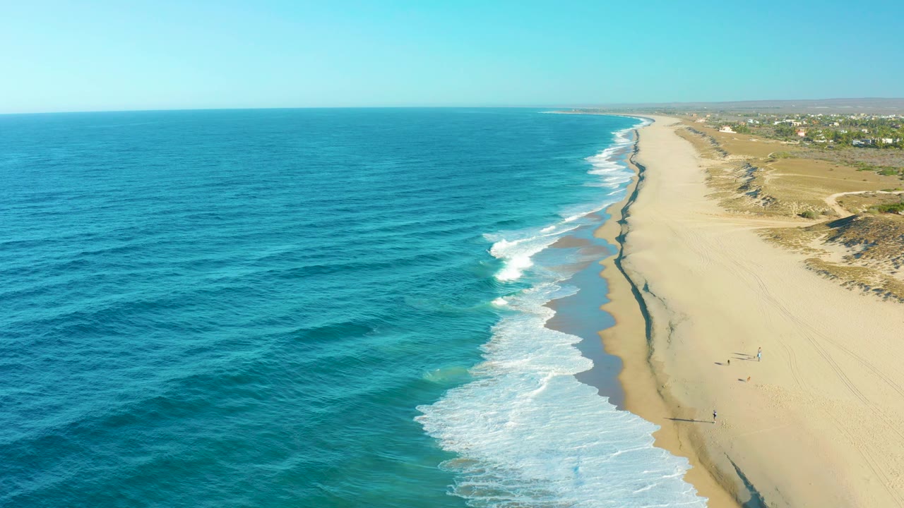 Drone-height view of Todos Santos surf curling diagonally across wet sand, with a few tiny people on the open beach and low dunes beyond.