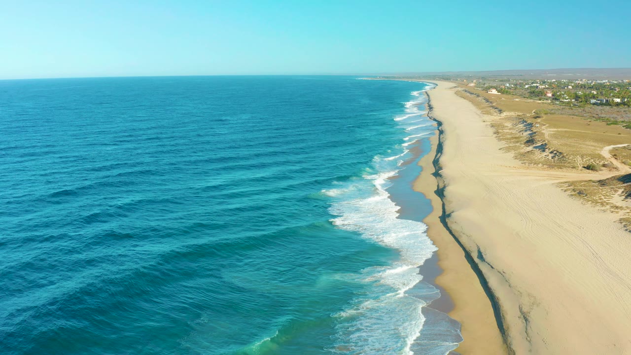 Aerial coastal frame in Todos Santos showing turquoise water, white wave bands, a long empty beach, dunes and sparse inland vegetation, with no visible people.
