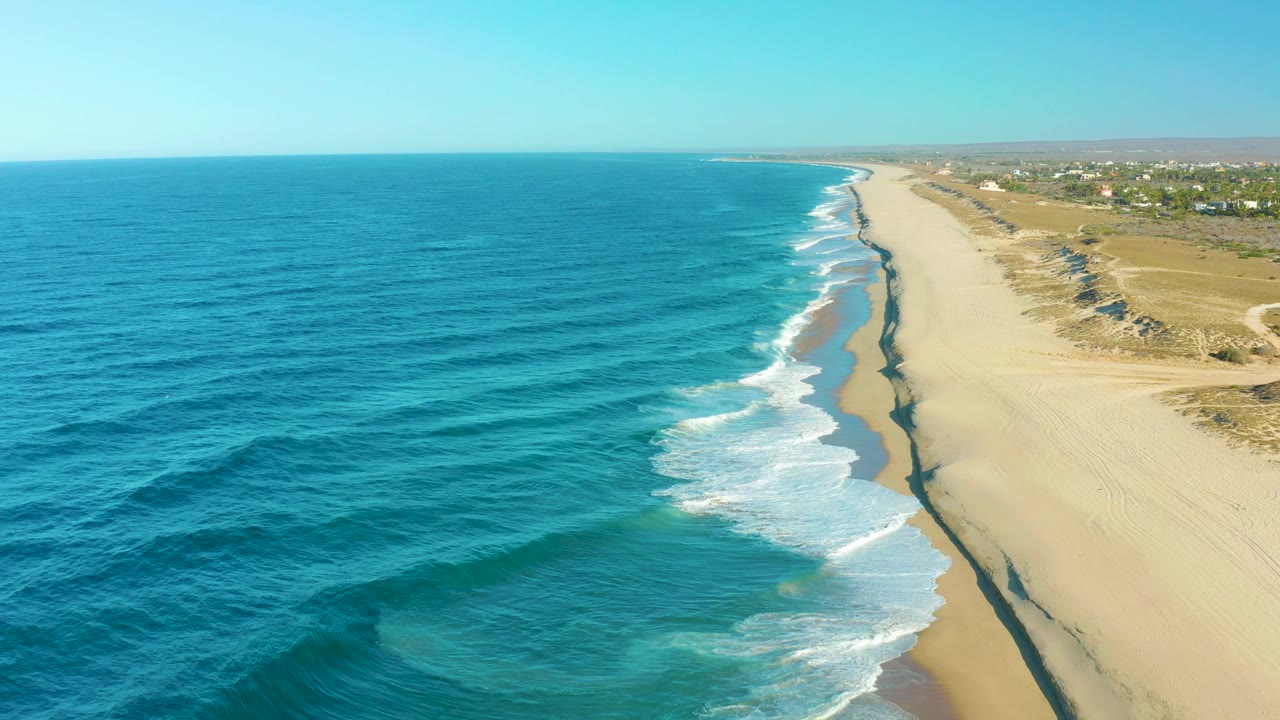Wide drone-style view over Todos Santos with ocean filling the left side, a pale sandy beach and surf line through the center, dry dunes to the right, and no visible people.