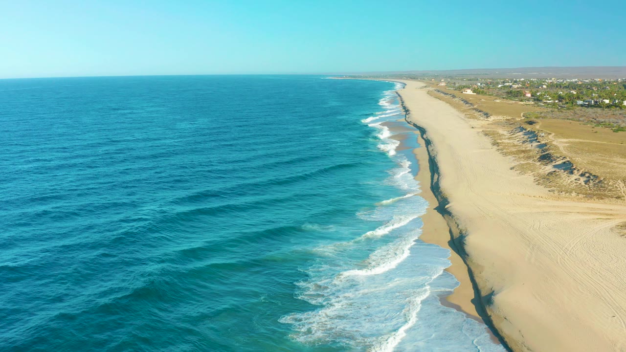 Aerial frame from Todos Santos with parallel wave lines moving toward pale sand, the shoreline running diagonally into the distance, inland dunes and buildings visible, and no visible people.