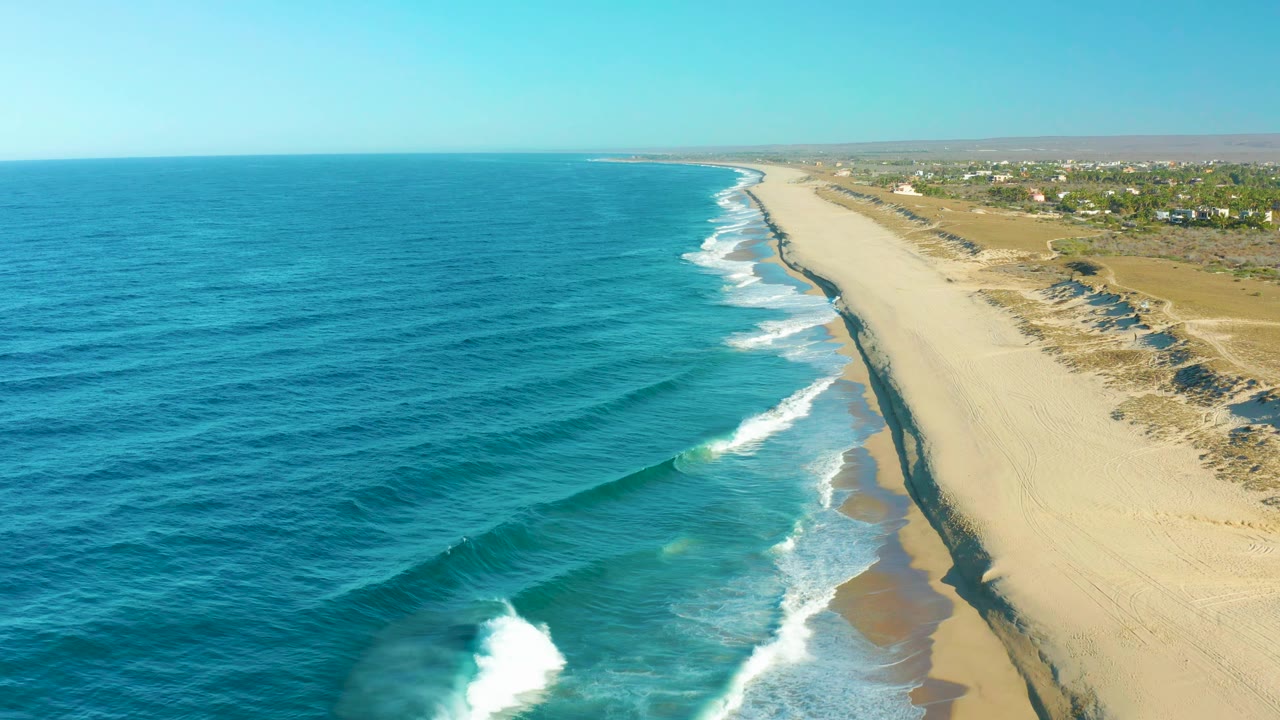 Aerial coastal frame in Todos Santos showing parallel blue wave bands, white foam near shore, a broad pale beach, dune ridges, and no visible people.