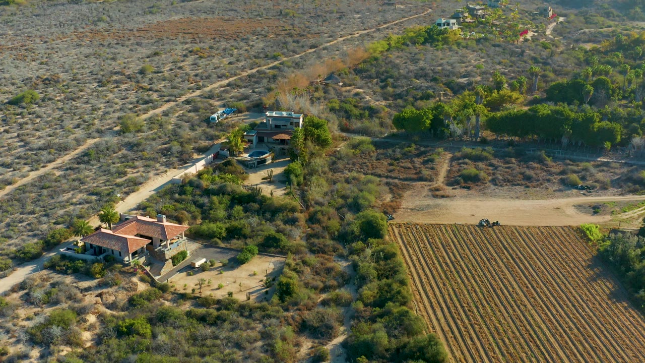 High aerial view in Todos Santos with the house lower left, the plowed field lower right, a small vehicle on the dirt road, and no visible people.