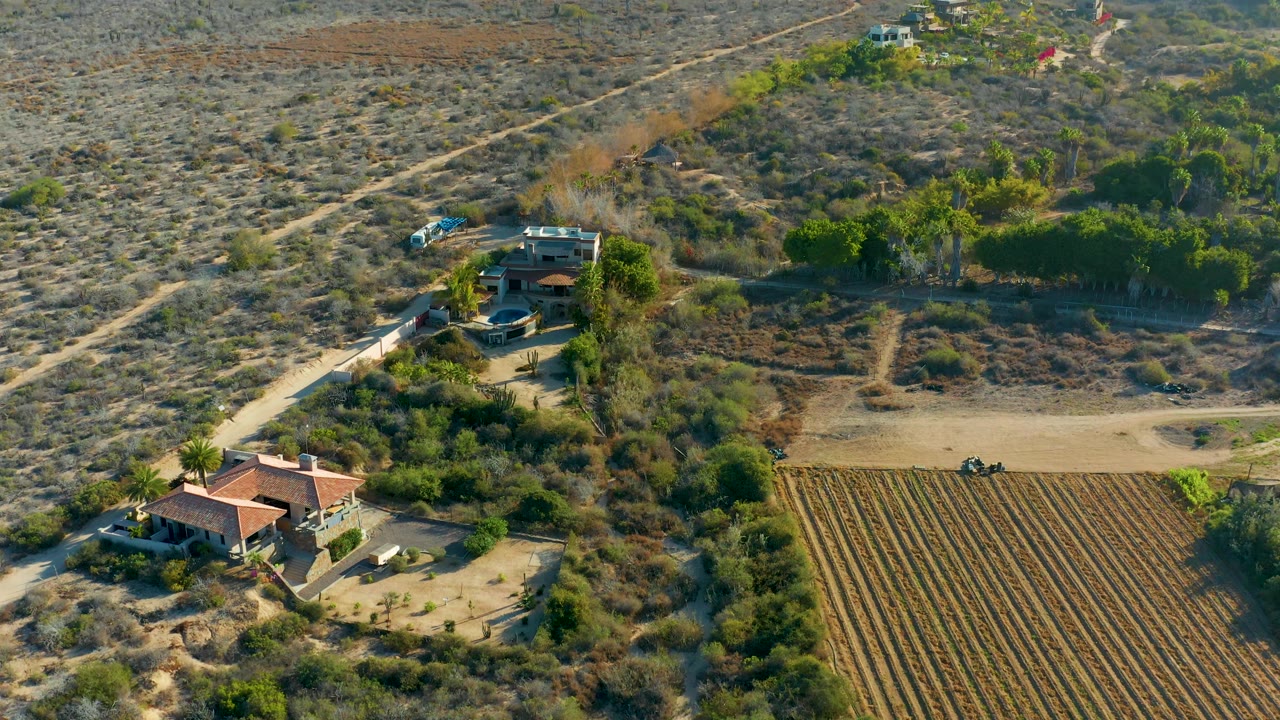 Oblique drone frame over Todos Santos showing scattered rural buildings, pale dirt tracks, a rectangular cultivated field, dry brush, and no visible people.