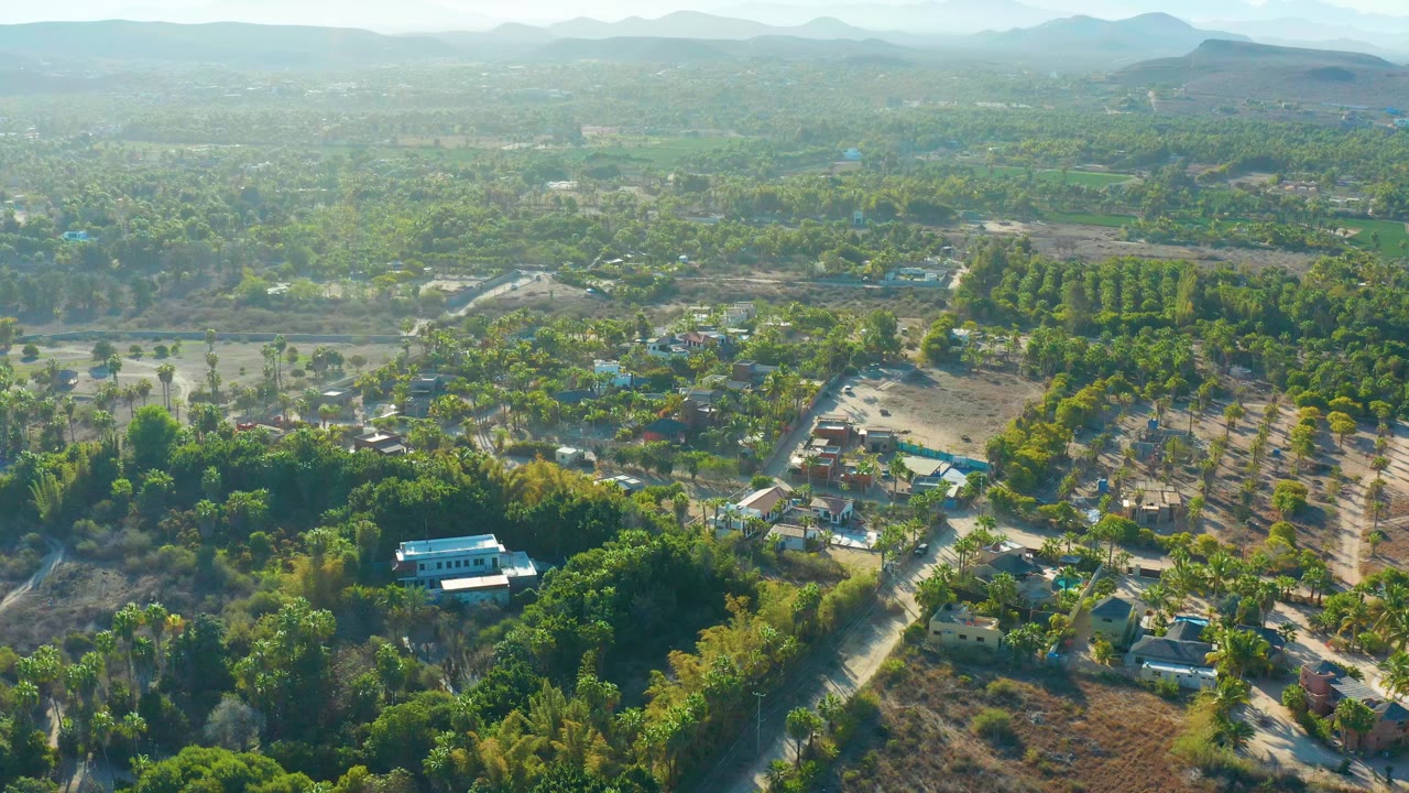 Opening aerial frame over rural Todos Santos, Mexico with scattered homes, pale roads, green tree cover, open fields, hazy mountains, and no visible people.
