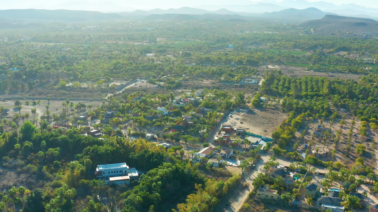 High aerial view across trees, fields, small buildings, and low mountains in the distance.