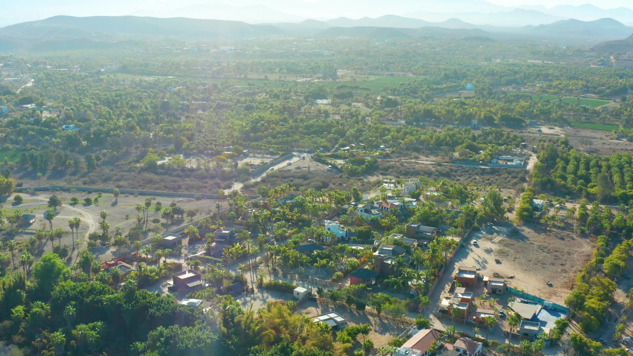 Aerial frame over Todos Santos showing a tree-filled residential area, pale roads, open ground, low buildings, distant mountains, and no visible people.