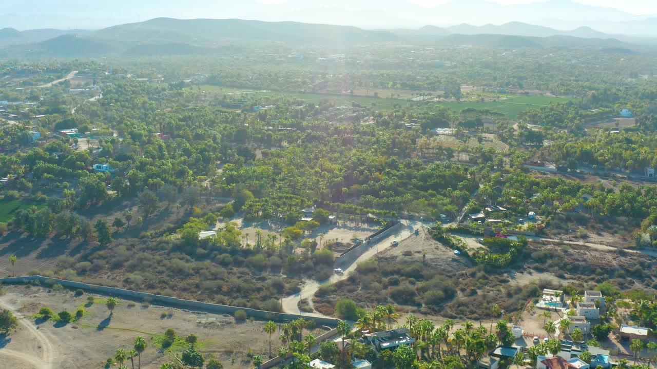 Wide aerial over Todos Santos, Baja California Sur, showing a road through dry scrub, green trees, scattered houses, hazy hills, and no visible people at the start of the clip.