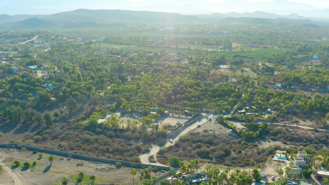 High aerial view over Todos Santos showing dense tree patches, a pale road cutting through arid ground, scattered houses near the lower edge, hazy mountains, and no visible people.