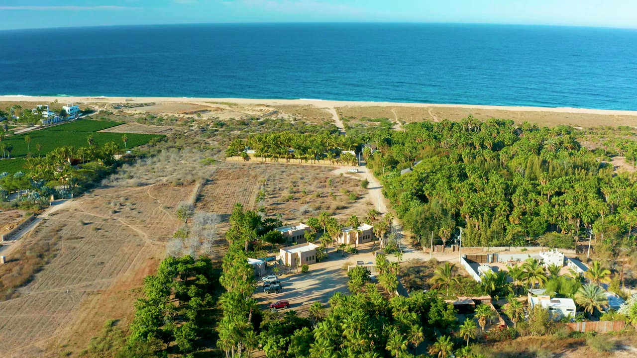Drone frame in Todos Santos, Baja California Sur with the ocean horizon above a beach strip, brown fields, tree clusters, small structures, dirt roads, and no visible people.