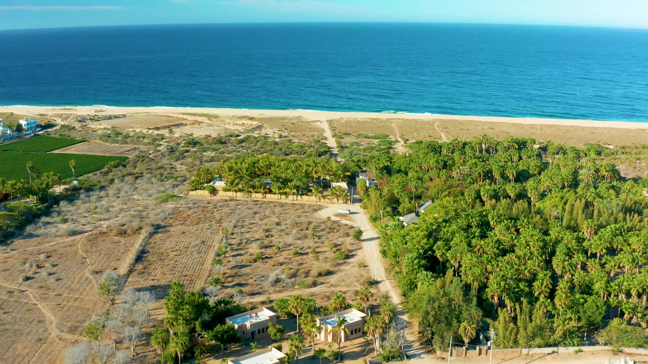 Aerial view in Todos Santos, Baja California Sur with the ocean horizon high in frame, pale beach below it, dry farmland at left, a dirt road through the center, and no visible people.