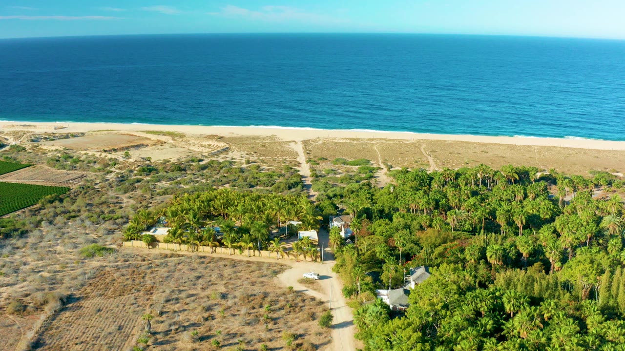 Aerial frame over Todos Santos, Mexico, with blue ocean beyond a pale beach, dry scrubland, green trees, dirt tracks, small structures, and no visible people.