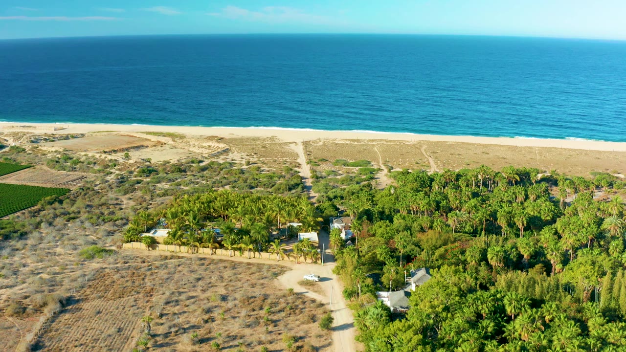 Wide aerial view of ocean, beach, dirt tracks, green vegetation, and a small white car.