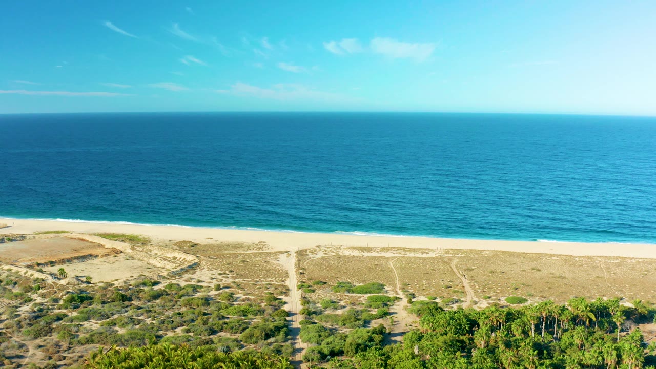 Mid-clip aerial frame in Todos Santos with the ocean horizon high across the image, pale beach below it, sandy dunes and paths in the foreground, and no visible people.