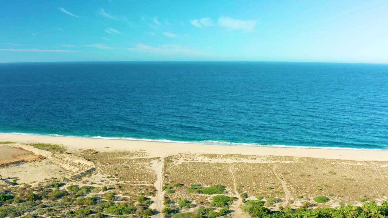 Todos Santos aerial frame with more open water across the upper view, a narrow empty beach below it, dune tracks, green scrub, and no visible people.