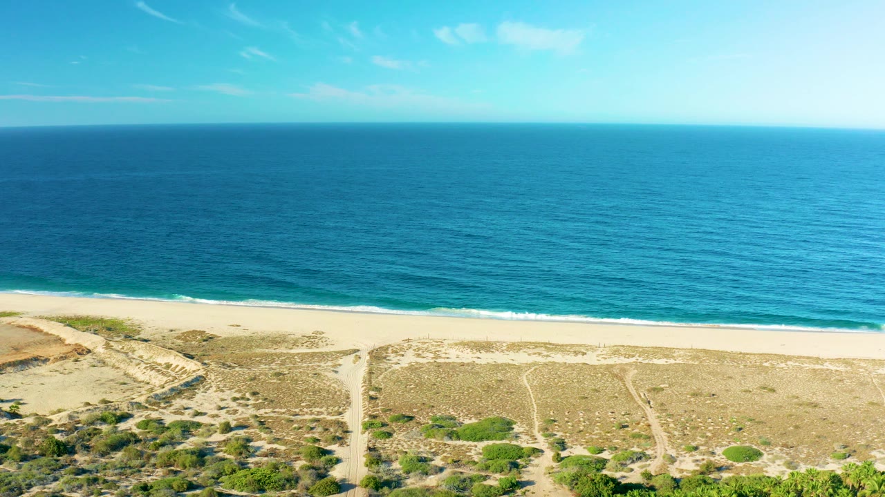A bright aerial view of the Todos Santos shore shows turquoise water near the beach, darker blue ocean beyond, sandy dunes, low vegetation, and no visible people.