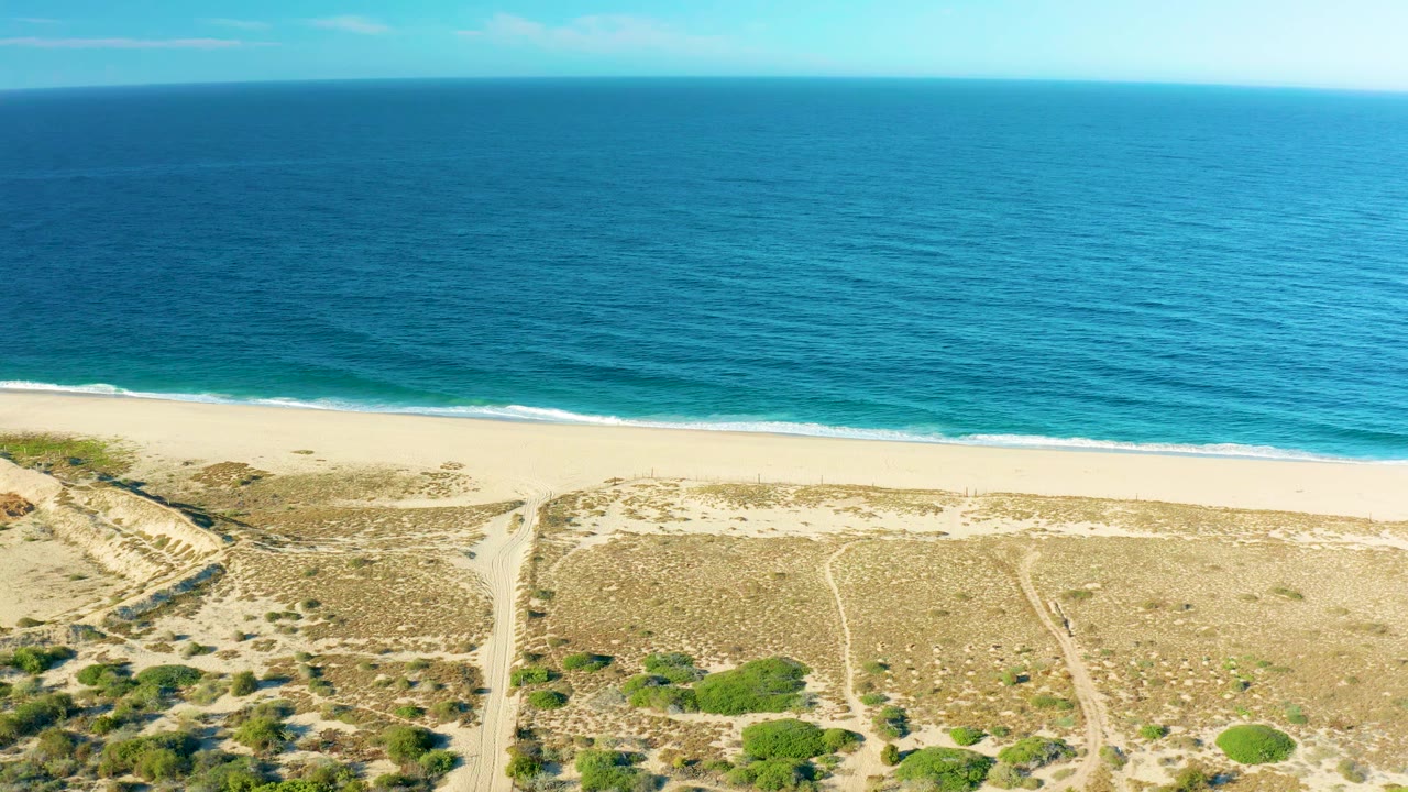 A wide aerial over Todos Santos shows the turquoise ocean, thin white waves, empty sand, and intersecting paths through low dune vegetation, with no visible people.