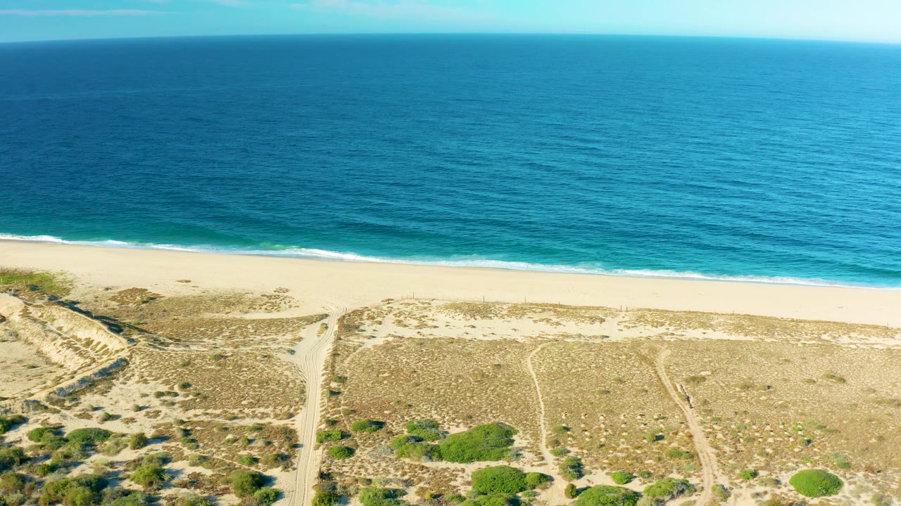 The frame holds a level ocean horizon above the Todos Santos shoreline, with bright water, a narrow beach band, and textured dune scrub below, with no visible people.