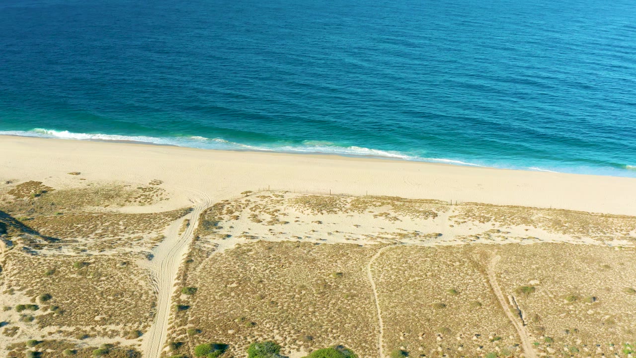 At 0:01, the Todos Santos shoreline remains framed from above as a thin white wave line reaches the sand, with dunes and faint paths below and no visible people.